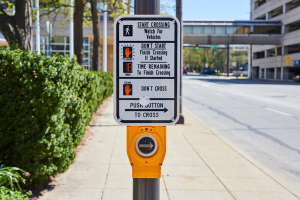 Pedestrian crossing signal button and instruction sign on a city sidewalk near an intersection.