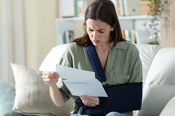 A woman with long brown hair wearing a green button-down shirt and an arm sling, looking concerned while reading medical bills.