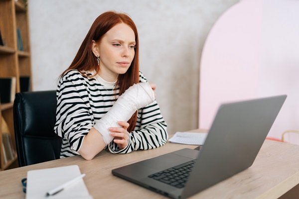 A woman with her arm in a white cast sits at a desk, looking focused and slightly frustrated while looking at an email from the insurance company on her laptop.